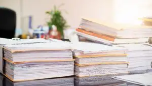 Stacks of documents and folders piled on an office desk, with a blurred background and sunlight streaming in. The scene conveys a busy, cluttered workspace.