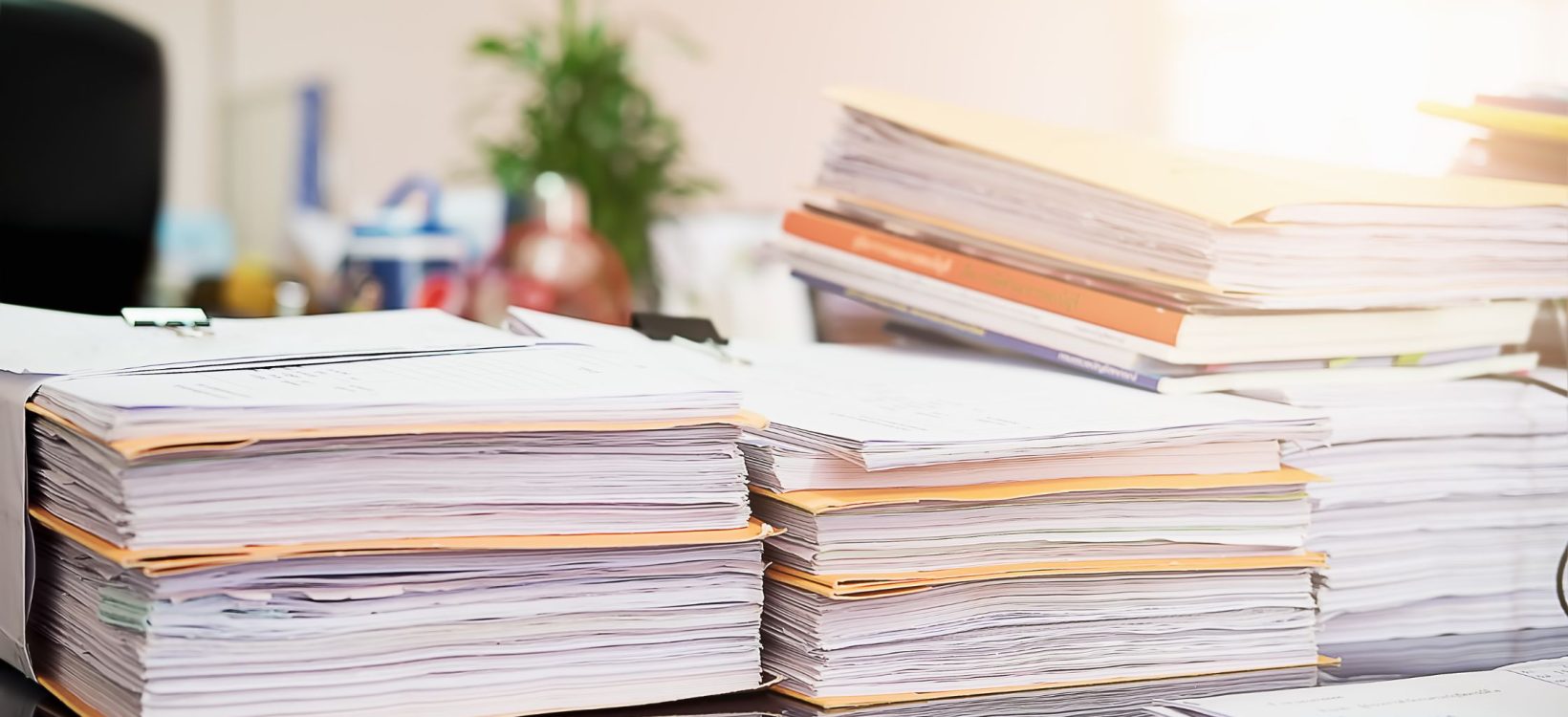 Stacks of documents and folders piled on an office desk, with a blurred background and sunlight streaming in. The scene conveys a busy, cluttered workspace.